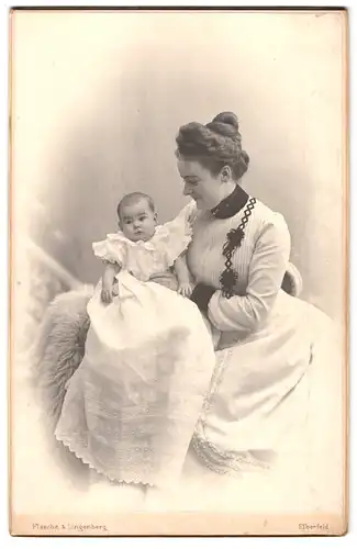 Fotografie Flasche & Klingenberg, Elberfeld, Frau in gestreiftem Kleid hält ihr neugeborenes Kind in weissem Kleidchen