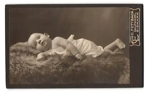 Fotografie C. Fettback, Salzwedel, Neuperverstrasse 28, Portrait niedliches Kleinkind im weissen Hemd auf Fell liegend