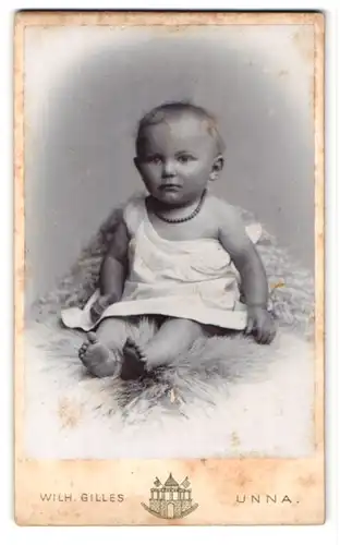 Fotografie Wilh. Gilles, Unna, Portrait niedliches Kleinkind im weissen Hemd auf Fell sitzend