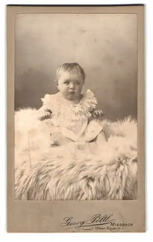 Fotografie Georg Pöltl, Miesbach / Ob.-Bayern, Portrait niedliches Kleinkind im weissen Kleid auf Fell sitzend