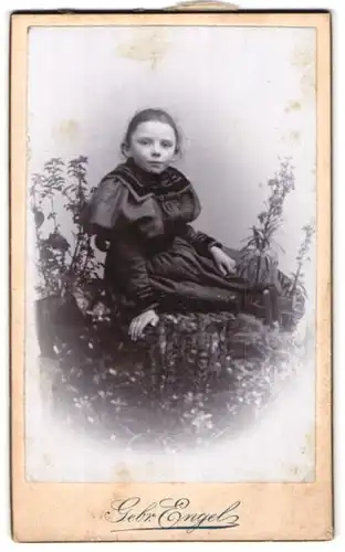 Fotografie Gebr. Engel, Diedenhofen, Portrait kleines Mädchen im schwarzen Kleid zwischen Blumen