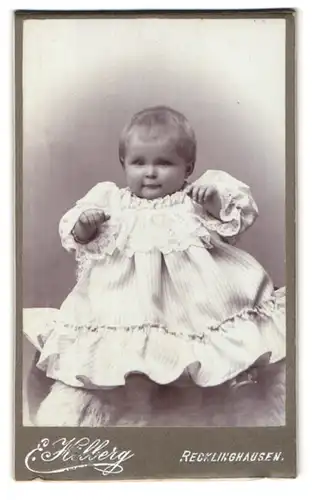 Fotografie Ernst Hilberg, Recklinghausen, Portrait niedliches Kleinkind im hübschen Kleid auf Fell sitzend