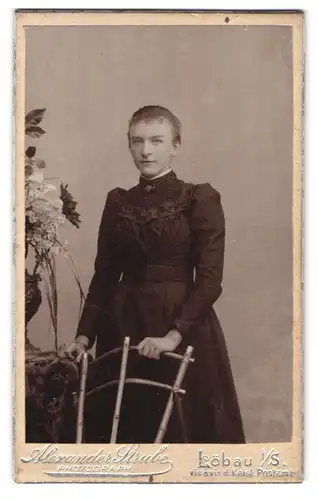 Fotografie Alexander Strube, Löbau i. Sa., Portrait charmant blickendes Fräulein im Kleid am Stuhl stehend