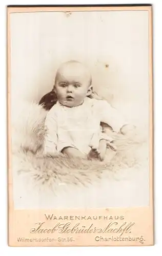 Fotografie Jacob Gebrüder Nachf., Berlin, Wilmersdorferstr. 56, Portrait süsses Baby im weissen Kleidchen auf Fell sitzend