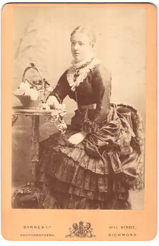 Fotografie Byrne & Co., Richmond, Hill Street, Portrait junge Dame im Kleid am Tisch sitzend