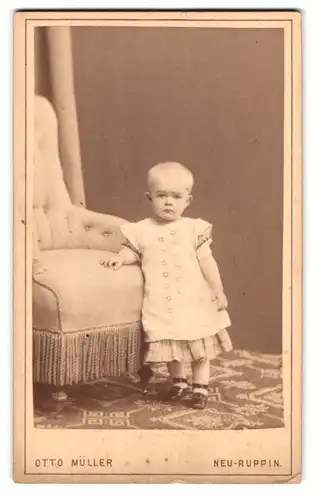 Fotografie Otto Müller, Neu-Ruppin, Portrait niedliches Kleinkind in hübscher Kleidung an Sessel gelehnt
