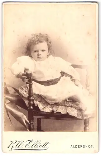 Fotografie R. W. Elliott, Aldershot, 93, High St., Portrait kleines Mädchen im weissen Kleid auf Stuhl sitzend