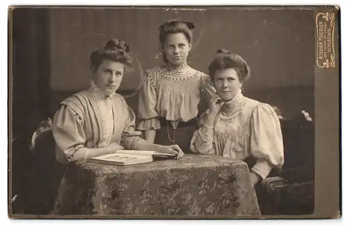 Fotografie Heinr. Petersen, Schleswig, Stadtweg 34, Portrait drei Junge Damen in hübscher Kleidung mit Buch am Tisch