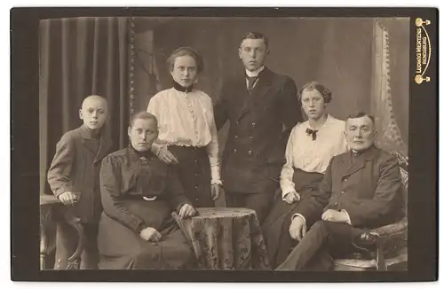 Fotografie Ludwig Mertens, Rendsburg, Portrait bürgerliche Familie in hübscher Kleidung am Tisch