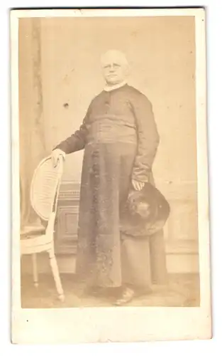 Fotografie B. Daems, Bruxelles, Gesitlicher in Gwand mit Bauchgurt hält Hut und trägt Brille