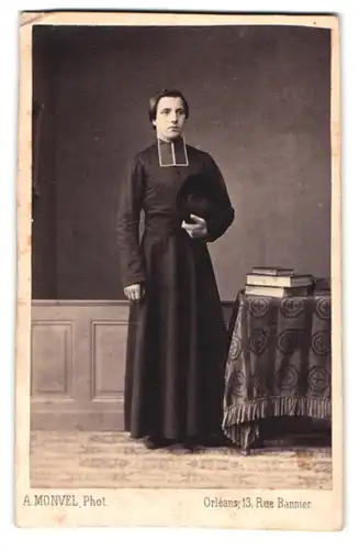 Fotografie A. Monvel, Orléans, Rue Bannier 13, Junger Geistlicher in Soutane hält einen Hut und steht neben Tisch