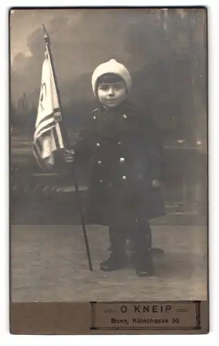 Fotografie O. Kneip, Bonn, Kölnstr. 50, Portrait niedlicher Bube mit Mütze im Wintermantel und Flagge in der Hand