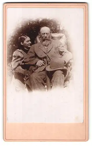 Fotografie unbekannter Fotograf und Ort, Älterer Herr mit Vollbart, Brille und Hut auf Sessel mit seiner Frau