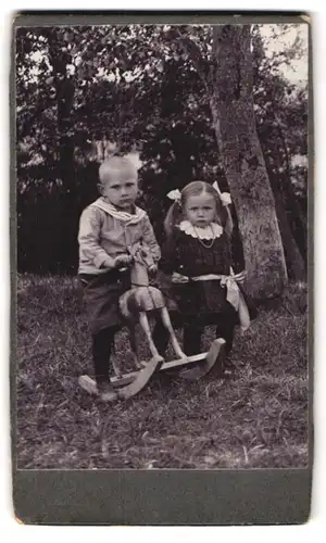 Fotografie Fotograf und Ort unbekannt, Portrait kleine Kinder mit Holzschaukelpferd im Garten