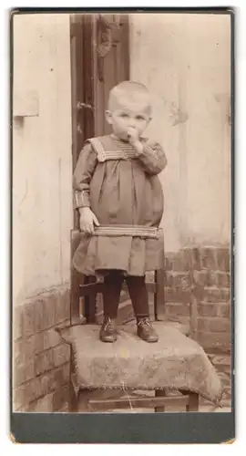 Fotografie Fotograf und Ort unbekannt, kleines Kind im Kleid steht auf einem Stuhl und lutscht am Daumen