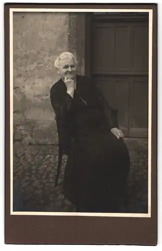 Fotografie unbekannter Fotograf und Ort, Portrait ältere Frau vor dem Eingang der Hauses sitzend