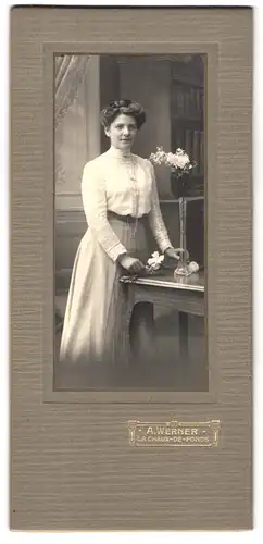 Fotografie A. Werner, La Chaux-de-Fonds, Hübsche Frau in langem Kleid mit hochgesteckter Frisur und Blumen in der Hand