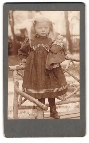 Fotografie unbekannter Fotograf und Ort, bildschönes Mädchen mit Puppe auf einer Holzbank stehend