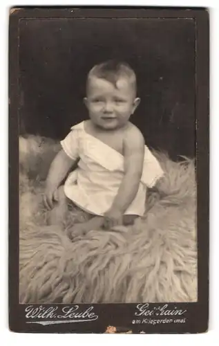 Fotografie Wilh. Leube, Geithain, Portrait süsses Kleinkind im weissen Hemdchen auf Fell sitzend