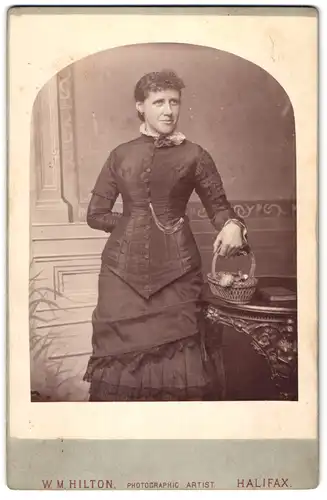 Fotografie W. M. Hilton, Halifax, 10 Corn Market, Portrait Dame mit Körbchen in gerafftem Kleid
