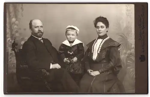 Fotografie Hofatelier Festge, Gera, Sitzender Mann mit Vollbart mit Frau und Kind in gepunktetem Mantel