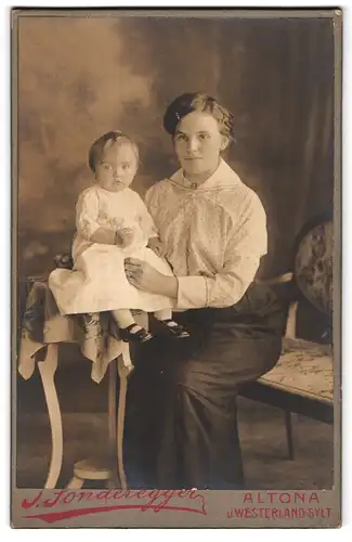 Fotografie J. Sonderegger, Hamburg - Altona, Gr. Bergstr. 15, Junge Frau in gepunktetem Kleid hält ihr Kleinkind in weiss
