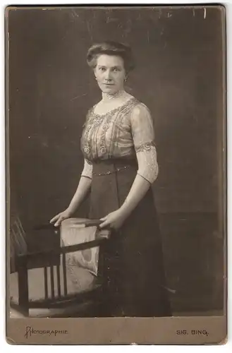 Fotografie Sig. Bing, Wien, Goldschmiedgasse 4, Dame mit bestickter Bluse trägt Ohrringe und steht hinter Stuhl
