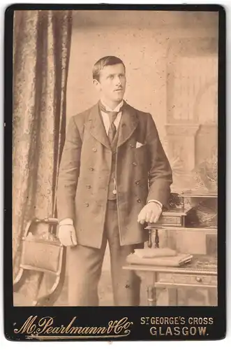 Fotografie M. Pearlmann & Co., Glasgow, St. George`s Cross, Portrait junger Mann im Anzug neben einem Mobilar