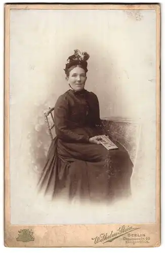 Fotografie Wilhelm Stein, Berlin, Chauseestr. 66, Frau mit verziertem Hut und Buch in schwarzem Kleid