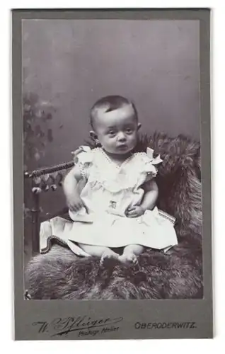 Fotografie W. Pflüger, Oberoderwitz, Portrait süsses Kleinkind im weissen Kleidchen auf Fell sitzend