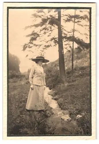 Fotografie unbekannter Fotograf und Ort, Frau im sommerlichen Kleid mit Hut an einem Bachlauf