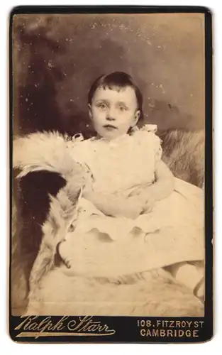 Fotografie Ralph Starr, Cambridge, 108 Fitzroy St., Portrait süsses kleines Mädchen im weissen Kleid auf Fell sitzend