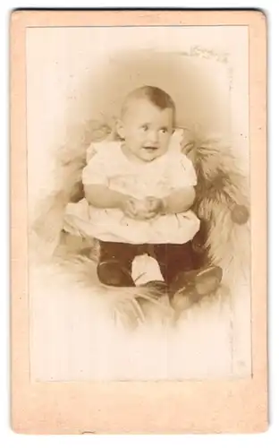 Fotografie unbekannter Fotograf und Ort, Baby im weissen Hemd mit Lächeln sitzend auf einem Fell
