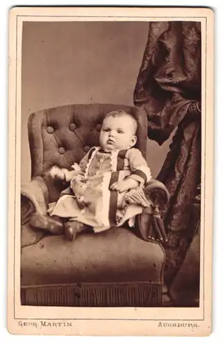 Fotografie Gebr. Martin, Augsburg, Bahnhofstr., Portrait süsses Baby im bestickten Kleidchen im Sessel lehnend