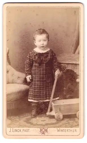 Fotografie J. Linck, Winterthur, Portrait kleines Mädchen im karierten Kleid mit einem Wagen