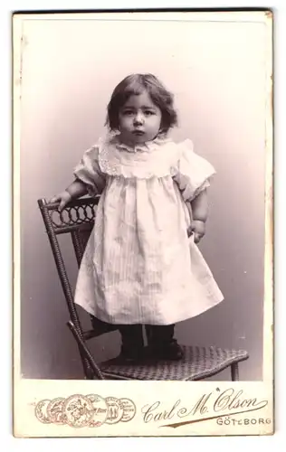 Fotografie Carl M. Olson, Göteborg, Masthuggstorget, niedliches Kleinkind in weissem Kleid auf Stuhl stehend