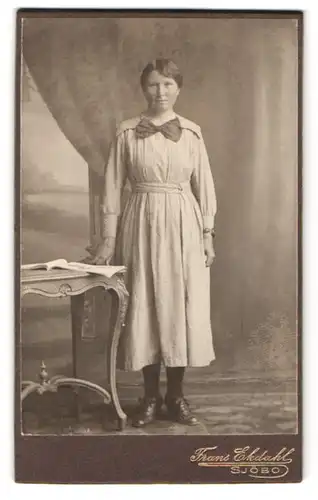 Fotografie Frans Ekdahl, Sjöbo, Rikstel 5, Portrait Frau im hellen Kleid mit Schleife im Atelier stehend