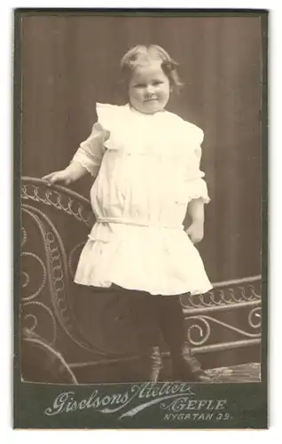 Fotografie Giselsons, Gefle, Nygatan 39, Portrait kleines Mädchen im weissen Kleid steht auf einer Bank