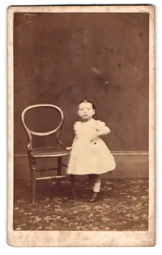 Fotografie Fotograf und Ort unbekannt, Portrait kleines Mädchen im Kleid neben einem Stuhl