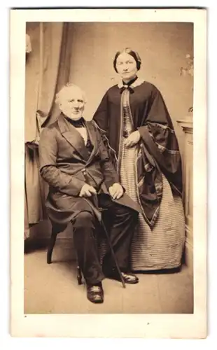 Fotografie Fotograf und ORt unbekannt, Portrait älteres Ehepaar in zeitgenössischer Tracht mit Stock