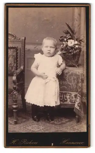 Fotografie H. Richers, Hannover, Cellerstr. 146, Portrait Kleinkind im weissen Kleid lehnt an einem Tisch