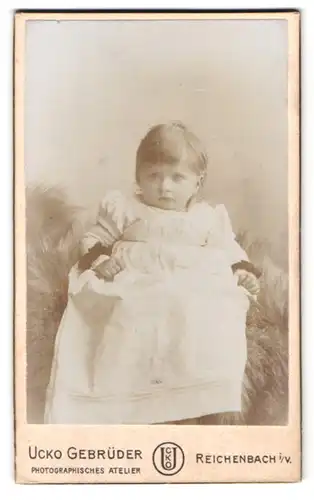 Fotografie Ucko Gebrüder, Reichenbach i. V., Portrait kleines Mädchen im weissen Kleid auf einem Fell sitzend