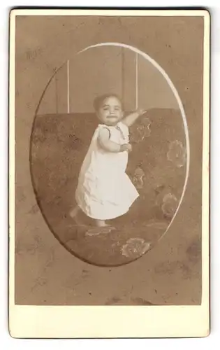 Fotografie Fotograf und Ort unbekannt, Portrait Kleinkind im Kleidchen auf einem Sofa