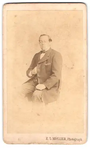 Fotografie E. L. Müller, Ort unbekannt, Portrait Mann im Anzug mit Fliege macht Napoleon Geste