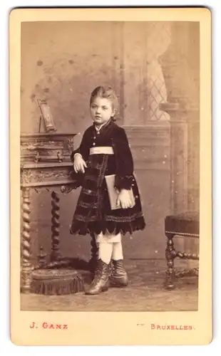 Fotografie J. Hanz, Bruxelles, Rue de l`Ecuyer 38, Portrait Mädchen im bestickten Kleid steht am Sekretär