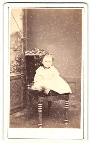 Fotografie Fotograf und Ort unbekannt, Portrait Mädchen im weissen Kleid sitzt auf einem Stuhl im Atelier