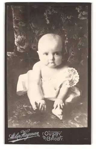 Fotografie Atelier Hagman, Broby, Niedlicher Säugling im weissen Kleid auf dem Sessel