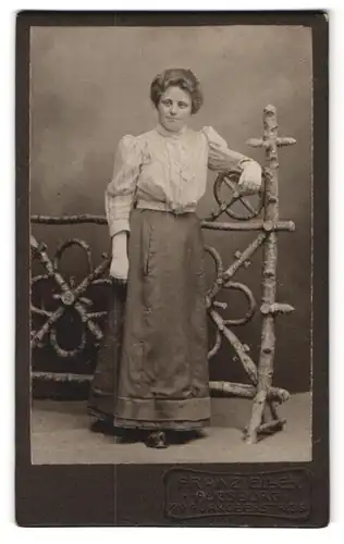 Fotografie Franz Eller, Augsburg, Jakoberstr. G. 3, Portrait Frau in gestreifter Bluse und Rock lehnt an einem Holzzaun