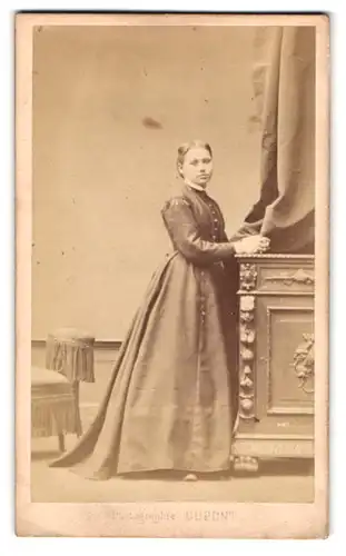 Fotografie Dupont, Bruxelles, Rue Neuve 67, Portrait junge Frau im Kleid lehnt an einer Kommode