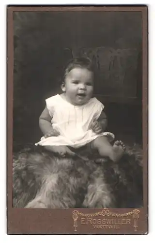 Fotografie E. Roggwiller, Wattwil, Portrait süsses kleines Mädchen im Kleidchen auf Fell sitzend